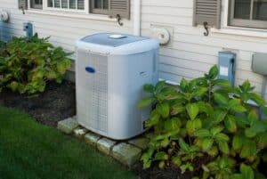 Air conditioning unit installed outside a house, surrounded by greenery and situated on a stone base.