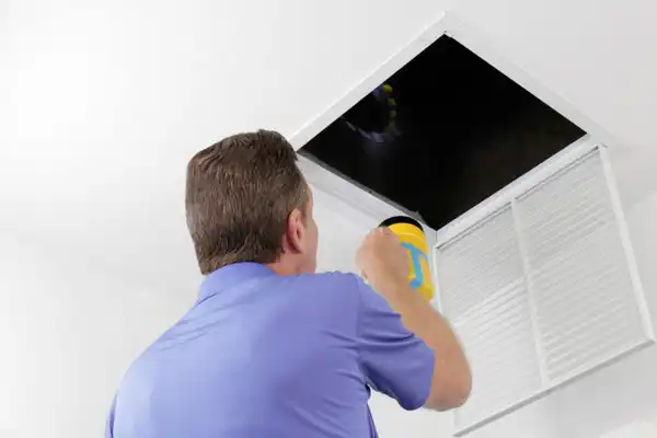 Person in a blue shirt shines a flashlight into a dark ceiling duct, standing near a white vent cover.