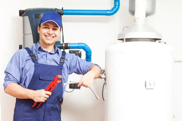 A plumber in blue overalls and cap holds a pipe wrench and a flexible pipe, standing next to a water heater with visible plumbing pipes in the background.