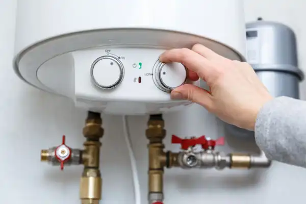 A hand adjusts the temperature control dial on a white water heater with visible pipes and valves below.