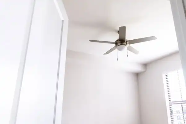 Ceiling fan with light fixture in a bright, empty room viewed from a doorway. White walls and a window with blinds are visible.