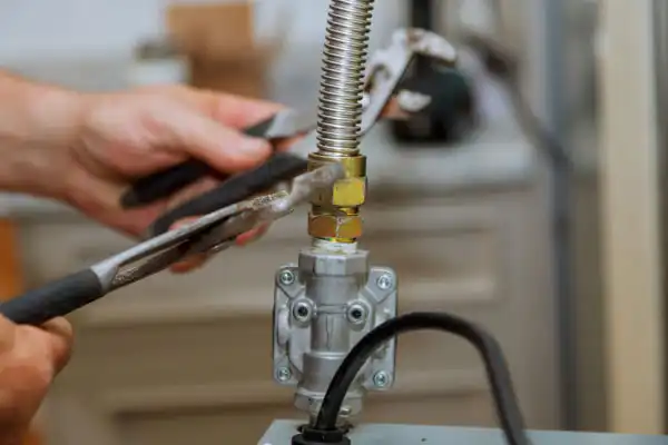 Person using pliers to tighten a metal gas connector on a gas line, with a background of an indoor setting.