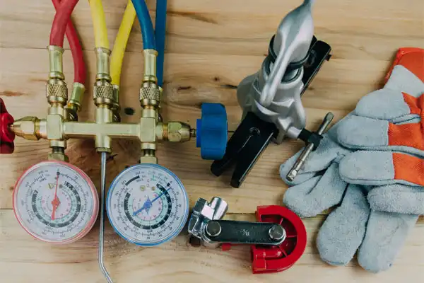 Tools for heating installation are spread across the wooden surface, featuring pressure gauges, colored hoses, a pipe cutter, a clamp, and work gloves.