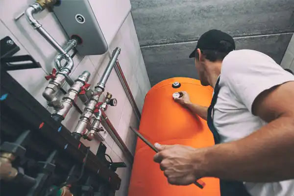 Plumber in cap and overalls expertly adjusts orange water heater in utility room with exposed pipes, showcasing his heating repair skills.