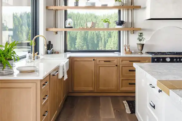 Bright kitchen with wooden cabinets, white countertops, and a large window. Open shelves display plants and dishes. Forest view outside.