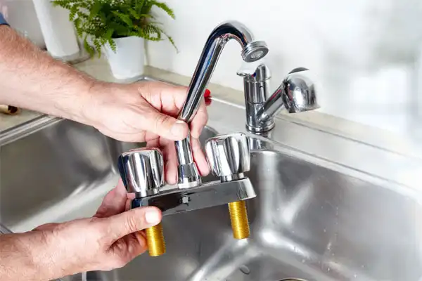 Person installing a new kitchen faucet over a stainless steel sink, with a potted plant in the background.