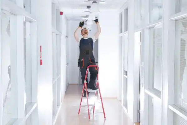 A person on a ladder is fixing a ceiling light in a bright hallway with glass-paneled doors.