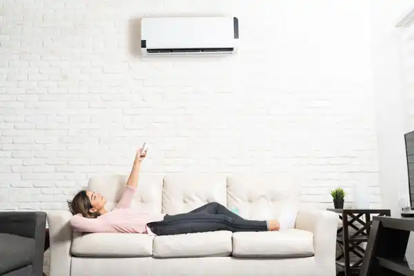 A person lounges on a white sofa, pointing a remote control at a wall-mounted air conditioner in a room with white brick walls.