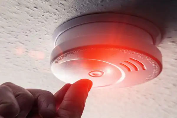 A person testing a smoke detector on the ceiling with a glowing red light indicating activation.