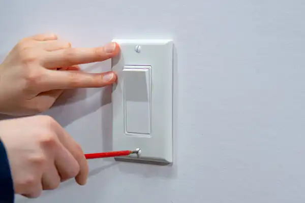 A person uses a red-handled screwdriver to tighten a screw on a white light switch cover while another hand holds the cover in place.