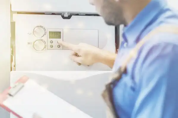 A person in a blue shirt adjusts the settings on a wall-mounted appliance, using the buttons on its digital display. They hold a clipboard in the other hand.