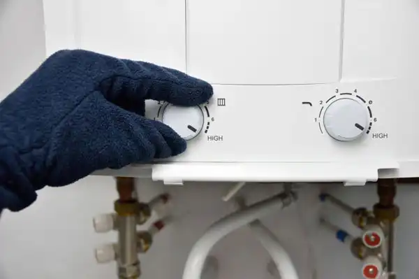 Gloved hand adjusts the temperature control knob on a water heater, with pipes visible below.