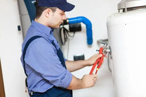 A plumber in a blue uniform and cap uses a wrench to work on a large white water heater, with pipes visible in the background.