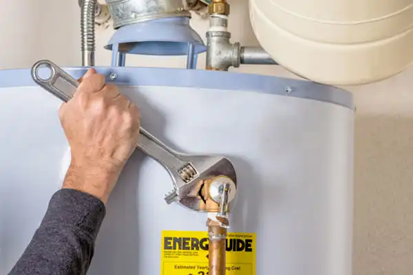 A person uses a wrench to tighten a pipe on a water heater. The heater has an energy guide label on it.