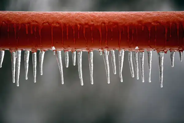 Red pipe with icicles hanging from the bottom against a blurred grey background.
