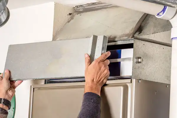 Hands removing the cover from a furnace filter compartment for maintenance in a residential setting.