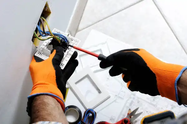 A person wearing orange and black gloves skillfully handles a screwdriver, focused on electrical repair as they install or fix an outlet. Nearby, tools and materials lie scattered, hinting at a dedicated work session.