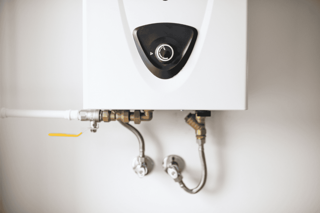 Close-up of a white tankless water heater mounted on a wall, showing control dial, pipes, and plumbing connections underneath.